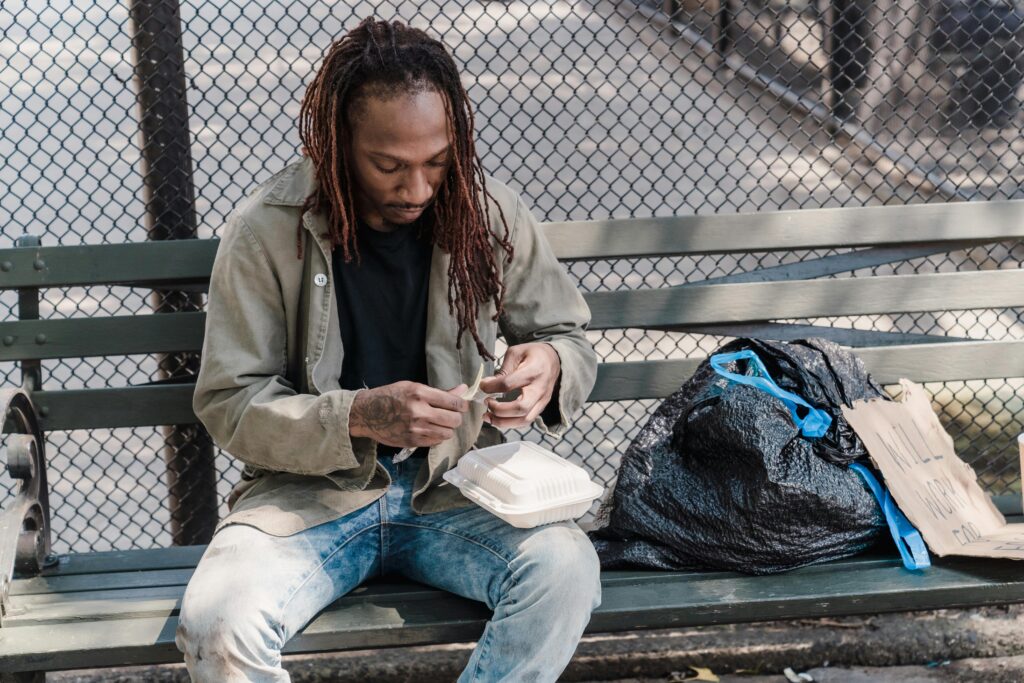 Young man with dreadlocks enjoying a meal from a container on a park bench outdoors.